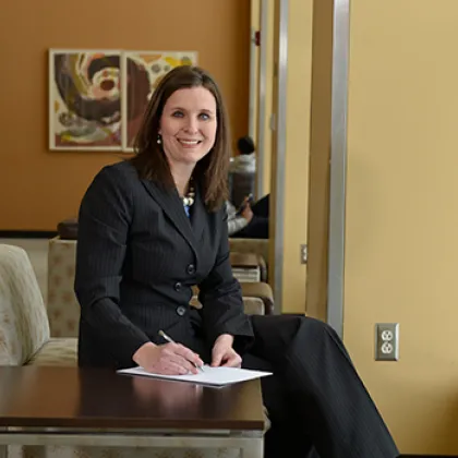 Rebekah Lewin sitting in a chair smiling at the camera. Her hand is poised over a notepad with a pen.